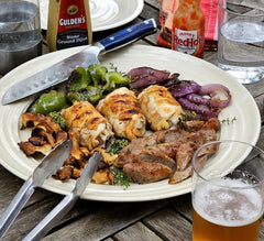 A plate of grilled meats and vegetables with condiments and a glass of beer on a wooden table, prepared on a SpitJack Tuscan Campfire Gridiron. All Stainless - 300 Sq. In. Grate.