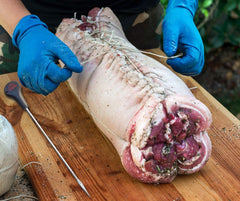 A person is preparing a large piece of meat on a cutting board using SpitJack's 12 Inch Trussing Needle and Butchers Cooking Twine Kit.