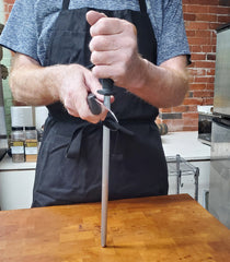 In a kitchen setting, someone is meticulously sharpening one of the SpitJack Barbecue Knife Bundle's knives on the included sharpening hone, ensuring it’s ready for the next culinary masterpiece.