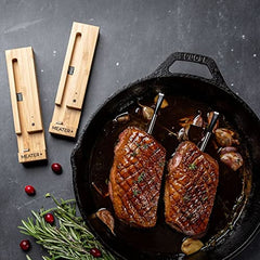 A cast iron skillet holds two cooked steaks with MEATER Plus: Dual Bundle Long Range Wireless Smart Meat Thermometers inserted. Two wooden thermometer holders from the MEATER brand are placed beside the skillet. Fresh herbs and cranberries are scattered around, showcasing both indoor and outdoor cooking versatility with wire-free cooking ease from MEATER.