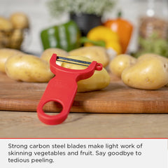 A red Kuhn Rikon Original Swiss Peeler with a robust carbon steel blade rests on a wooden surface, surrounded by potatoes and other vegetables in the background.