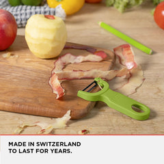 A green peeler from the Kuhn Rikon Original Swiss Peeler 3-Pack (includes Red/Green/Yellow) rests on a wooden cutting board with a partially peeled apple and apple peels. Various other fruits and vegetables appear in the background. Text reads: 