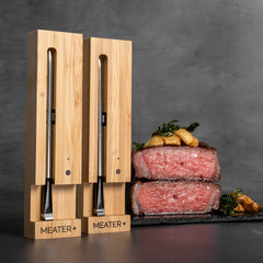 Two MEATER Plus: Dual Bundle thermometers stand upright next to a sliced medium-rare steak garnished with herbs and garlic on a dark serving board, perfect for wire-free cooking both indoors and outdoors.