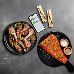 Two plates of cooked meats: one with lamb chops garnished with herbs, and one with a seasoned salmon fillet. An iPhone displaying a cooking app and two MEATER Plus thermometers from the MEATER brand, offering long-range wireless monitoring for hassle-free cooking, are beside the plates.