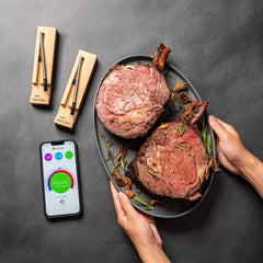 Person holding a plate of two cooked ribeye steaks, with a smartphone displaying a meat cooking app and two MEATER Plus: Dual Bundle Long Range Wireless Smart Meat Thermometers placed on the table, perfect for both indoor and outdoor cooking.