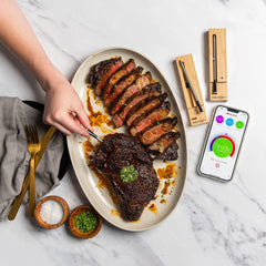 A person places the MEATER Plus: Dual Bundle, a long-range wireless smart meat thermometer from MEATER, into a sliced beef steak on a white platter, next to a mobile phone displaying the meat's temperature and readiness. Salt, chopped herbs, and utensils are nearby, making it perfect for both indoor and outdoor cooking in the oven, grill, kitchen, BBQ smoker, or rotisserie.