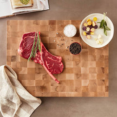 A raw tomahawk steak with sprigs of rosemary, a bowl of salt, a bowl of peppercorns, and a plate of assorted vegetables rest on a John Boos Boos Block CCB Series Large Reversible Wood Chopping Board in Maple beside a striped cloth.