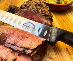 Close-up of a sliced roast beef with a knife from the SpitJack Barbecue Knife Bundle partially inserted, resting on a wooden cutting board, with a dish of vegetables in the background.