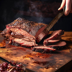 A hand slices smoked brisket on a wooden board with the SpitJack Deluxe Brisket Slicing Knife (11