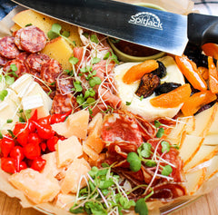 An assortment of cheeses, cured meats, dried fruits, and microgreens sits next to a large SpitJack barbecue knife from the bundle that includes four knives, a sharpening hone, and a case.