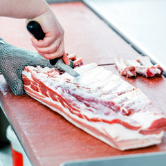 A person wearing a mesh glove is skillfully slicing a slab of raw meat with a knife from the SpitJack Barbecue Knife Bundle on a red cutting board.