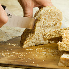 A person slices a loaf of sesame seed bread with a SpitJack BRSKT 2.0S Brisket Slicing Knife by SpitJack on a wooden cutting board.