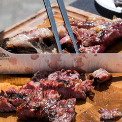 A juicy grilled steak with grill marks is sliced on a wooden cutting board using the SpitJack BRSKT 2.0S Brisket Slicing Knife, while tongs hold the seasoned meat in place.