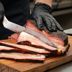 A person wearing black gloves slices smoked brisket on a wooden cutting board with the SpitJack BRSKT 2.0S Brisket Slicing Knife featuring a Granton edge.