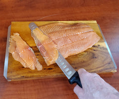 A hand expertly slicing thin pieces of salmon on a wooden cutting board using the SpitJack Barbecue Knife from the comprehensive Barbecue Knife Bundle, which includes 4 knives, a sharpening hone, and a case.