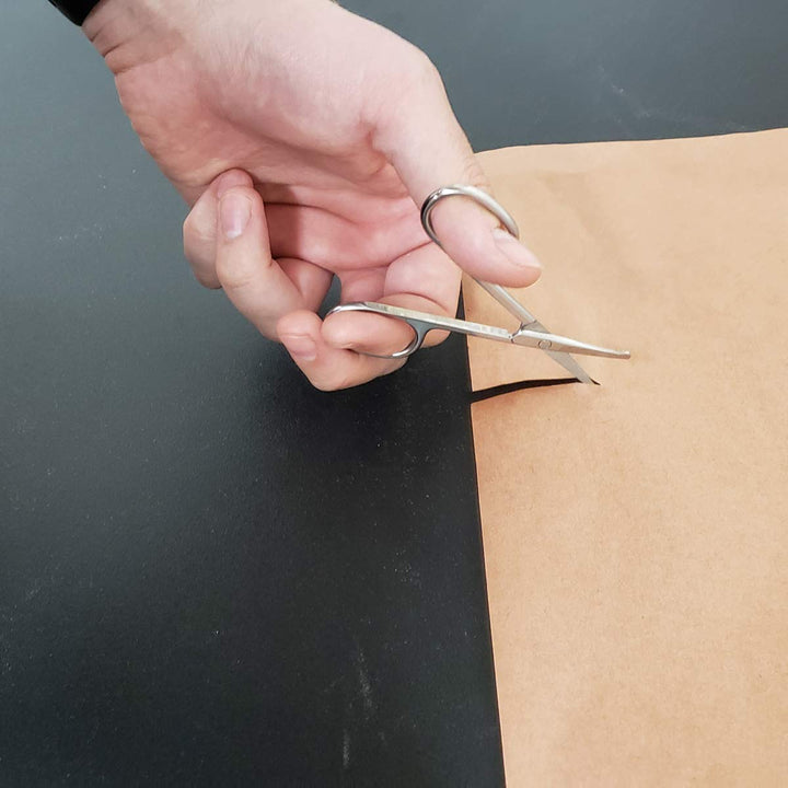 A hand is cutting a piece of brown paper with the small SpitJack Utility Mini Scissors (4 Inch Stainless Steel) from a 2 pack over a dark surface.