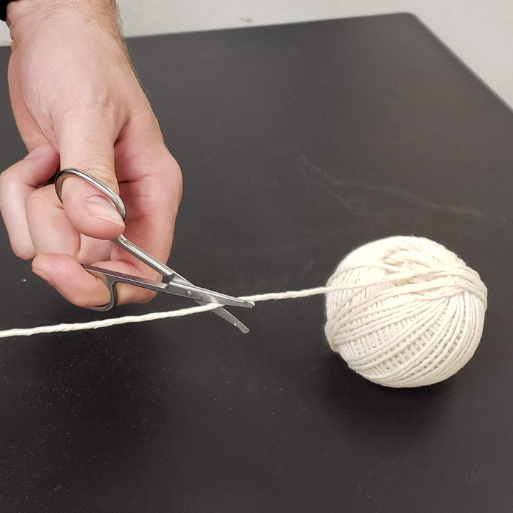 A hand holding a pair of SpitJack Utility Mini Scissors (4 Inch Stainless Steel) from the 2-pack cuts a piece of white yarn from a ball of yarn on a dark surface.