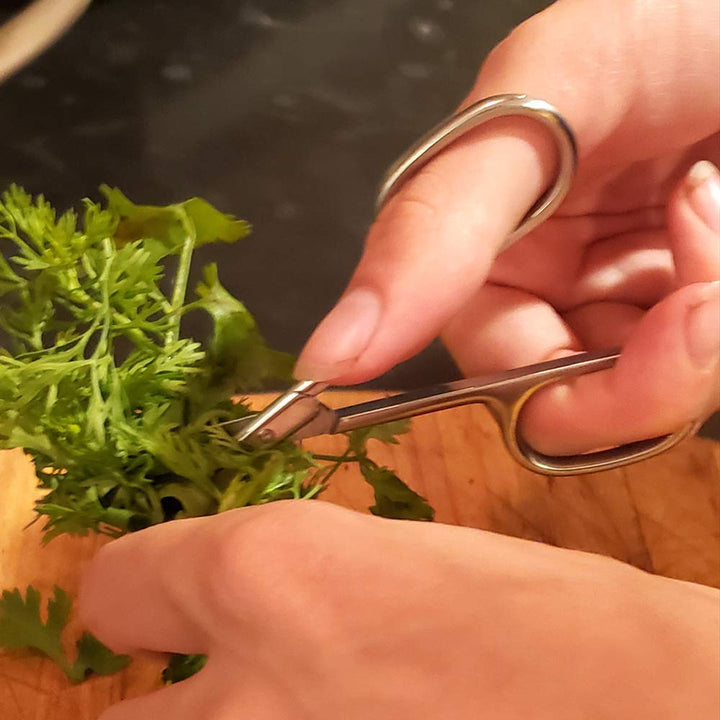 A person using SpitJack Utility Mini Scissors (4 Inch Stainless Steel) 2 Pack to cut fresh herbs on a wooden cutting board.