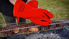 A person wearing SpitJack Deluxe Fireplace - Barbecue Gloves FP is grilling meat on a grill.