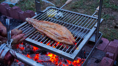 A person grilling a fish on a SpitJack ALL Stainless Tuscan Fireplace and Camping Grill Bundle - DX grill.