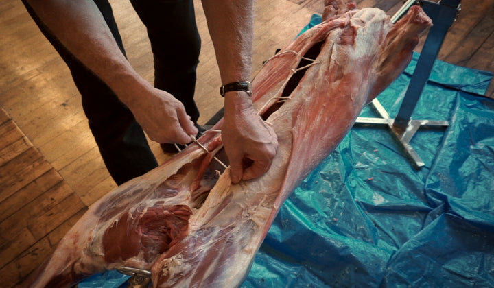 A man cutting a piece of meat on a table using the SpitJack 7" Stainless Steel Meat Trussing Needle and Butcher&