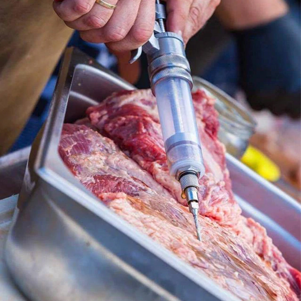 A person uses the SpitJack MAGNUM Meat Injector Complete Kit with Deluxe Case and Four Needles to inject marinade into raw meat over a stainless steel tray—ideal for BBQ competitors.