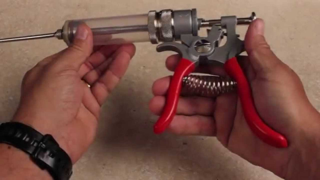 A person holds The SpitJack MAGNUM Meat Injector—complete kit with a clear barrel, metal needle, red handles, and deluxe case—against a beige background. This trusted tool comes with four needles and is favored by BBQ competitors.